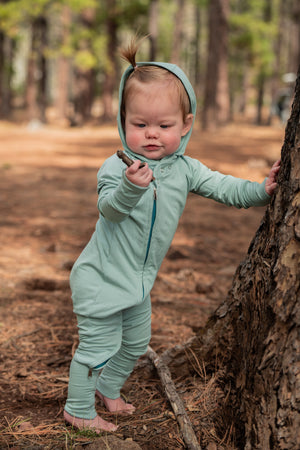 Child in a romper with a hood standing next to a tree in a forest