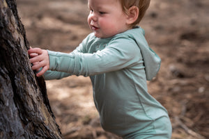 Child in a light hoodie romper standing next to a tree in a natural setting feeling the bark