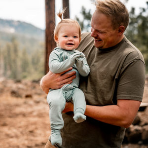 Man holding a baby outdoors with trees and mountains in the background, romper covers baby's hands and feet