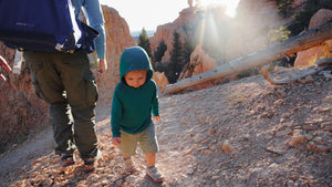 Child in a green hoodie walking on a rocky trail with a person and scenic landscape.