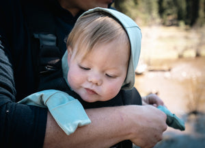 Baby in a light blue hood being held outdoors with a blurred natural background cuffs of romper is covering her hands